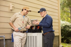 two-smiling-ac-technicians-standing-over-ac-outdoor-unit-looking-at-mobile-tablet