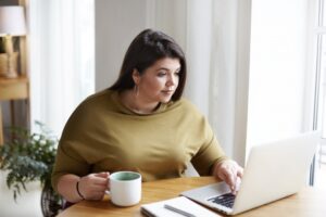 plus-sized-woman-looking-at-laptop-drinking-coffee-at-desk